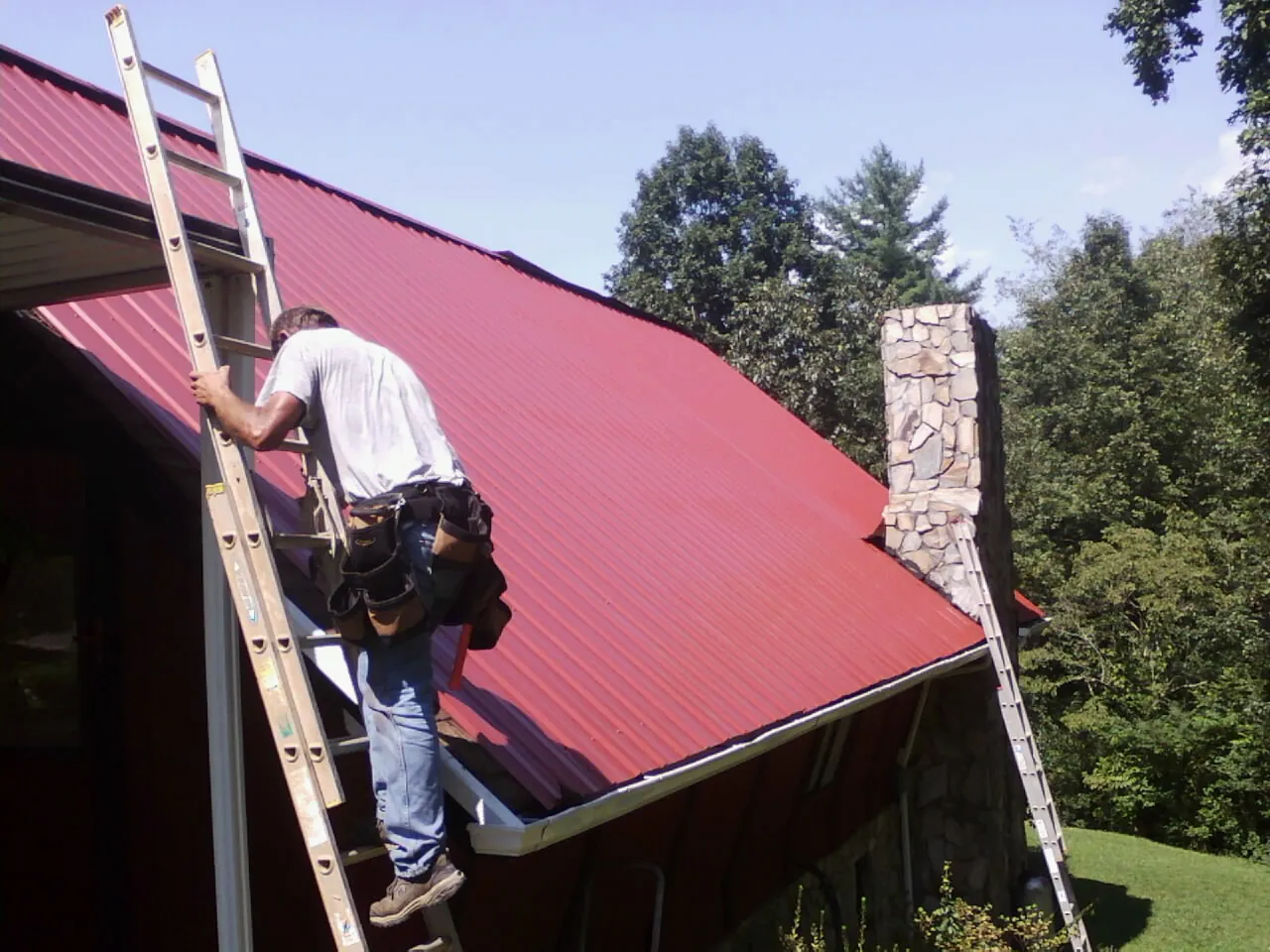Roofer on a ladder beside a red metal roof and stone chimney — Soffit & Fascia Repair by Roofing Brothers in East Brady